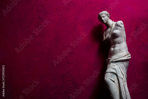 Vintage plaster statue, Aphrodite of Melos or Venus de Milo, wearing a beaded necklace, on dark pink weathered background. High-contrast photo with copyspace
