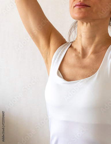 Shot of the woman in the white top against the white wall, performing self examination of the breasts, looking for abnormalities. Breast cancer awareness