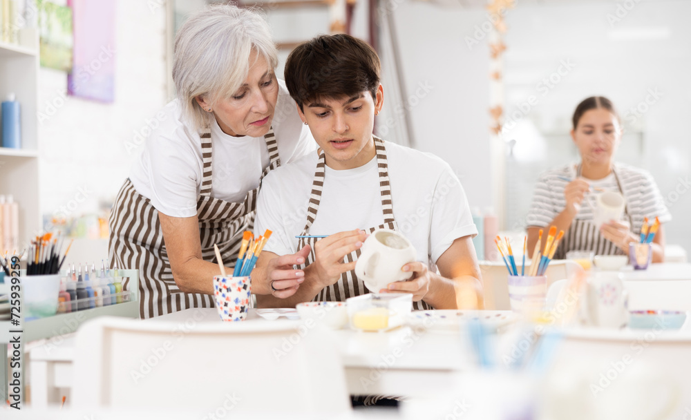 Fototapeta premium Passionate elderly female teacher offering guidance to interested young guy painting ceramic mug in pottery class. Creative hobby concept..