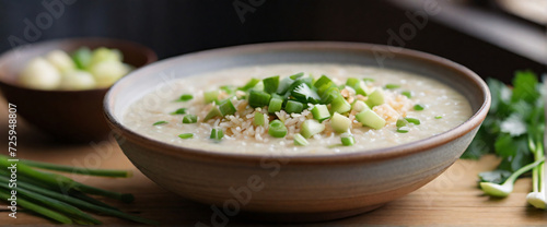 A steaming bowl of rice porridge, placed centrally on a textured, earthenware dish. The porridge is garnished with a sprinkle of chopped spring onions, a few slices of ginger, and a light drizzle