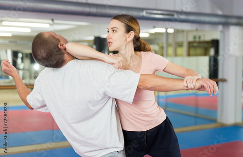 Concentrated sporty young woman learning self defence techniques in sparring with man, practicing elbow blow with wristlock to opponent in gym