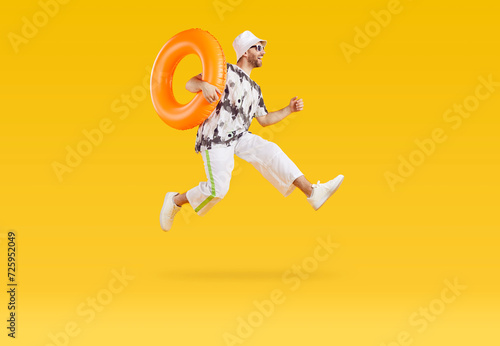 Excited young man jumping with inflatable rubber ring. Full body length portrait of happy man in sunglasses, casual summer clothes and panama hat having fun on isolated yellow studio background
