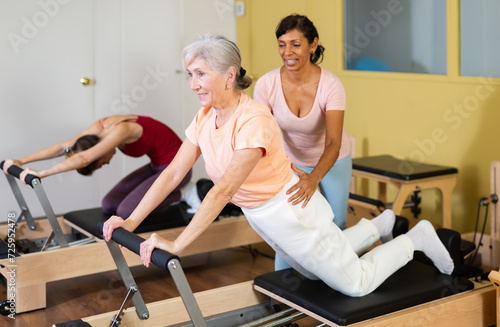 Aged woman doing pilates on reformer in fitness studio with Hispanic female personal trainer controlling movements