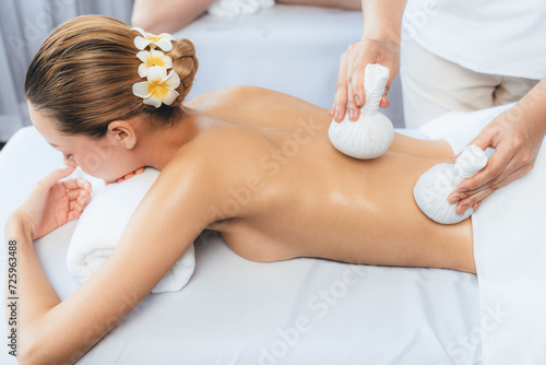 Hot herbal ball spa massage body treatment, masseur gently compresses herb bag on couple customer body. Tranquil and serenity of aromatherapy recreation in day lighting ambient at spa salon. Quiescent