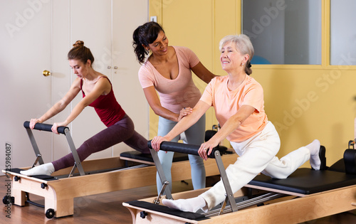Two active women practicing Pilates in a fitness studio perform an exercise using a reformer bed, where a female instructor ..helps them do it correctly