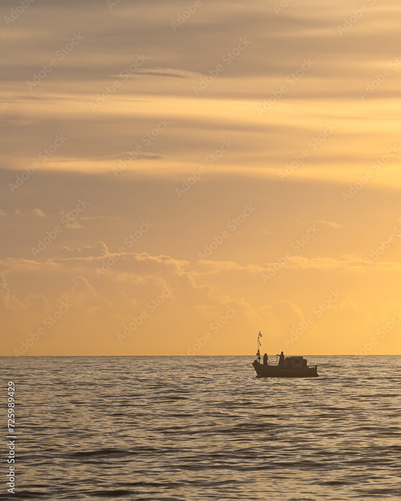 A boat sailing on the sea with clouds and the horizon in the background. Silhouette of a fishing boat sailing alone in the vastness of the sea at sunrise.