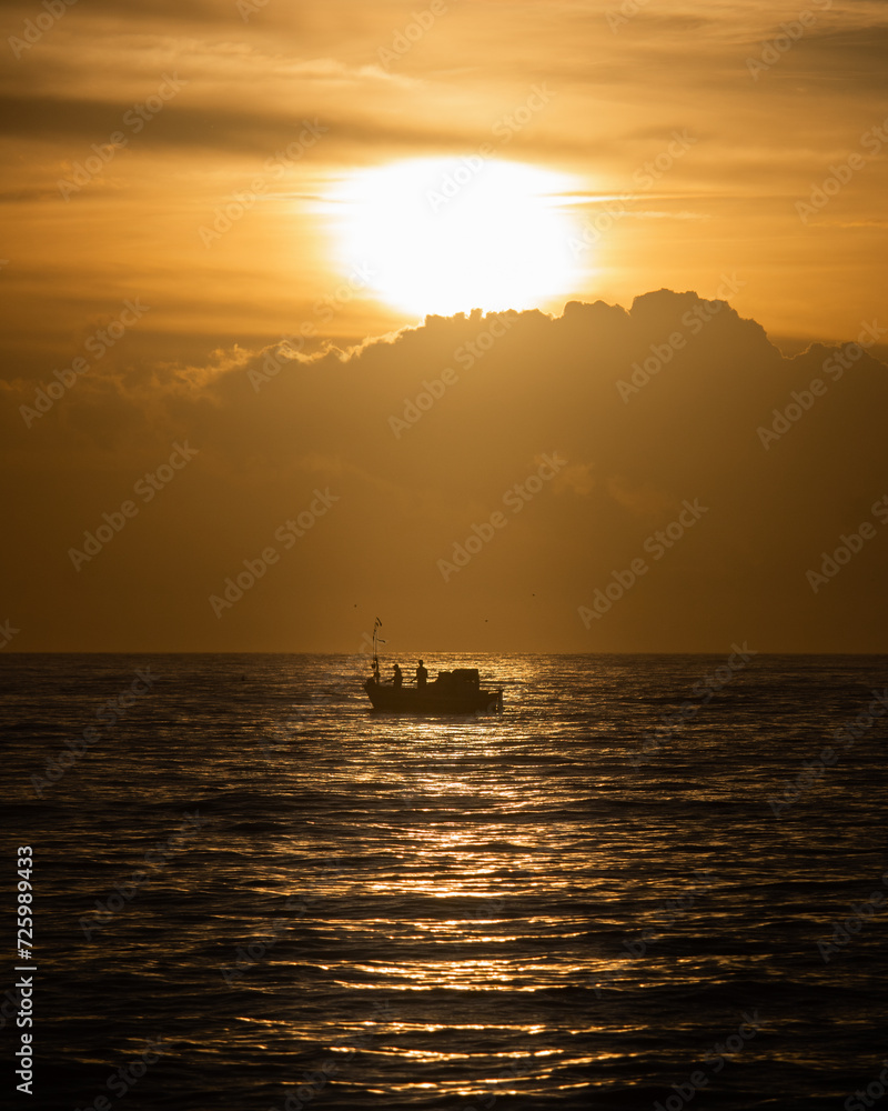 A boat sailing on the sea with clouds and the horizon in the background, under the sunrise. Silhouette of a fishing boat sailing alone in the vastness of the sea at sunset.





