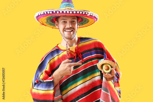 Happy young Mexican man in sombrero and with avocado on yellow background