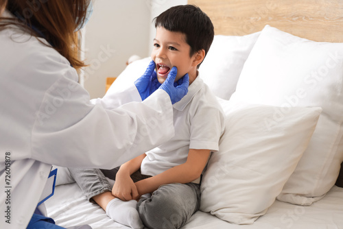 Ill little Asian boy showing sore throat to doctor in bedroom