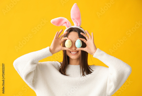 Funny young woman with bunny ears and Easter eggs on yellow background