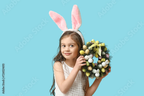Cute little girl in bunny ears with Easter wreath on blue background