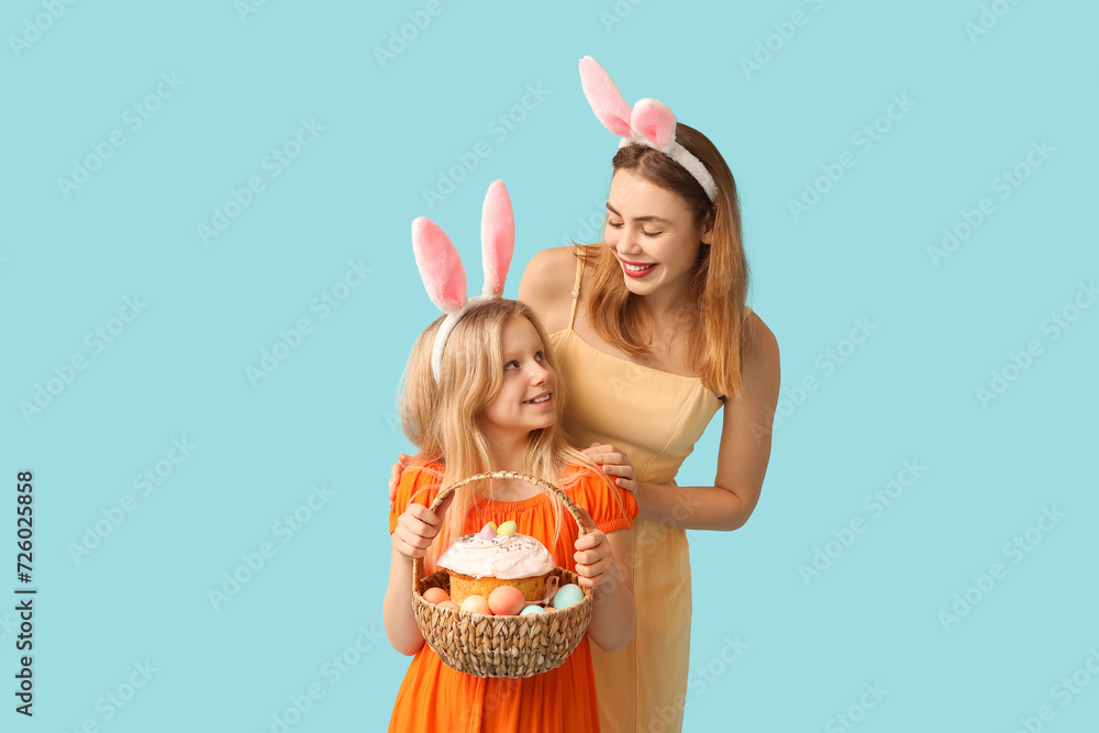 Young woman and her daughter in bunny ears holding basket of Easter cake with painted eggs on blue background