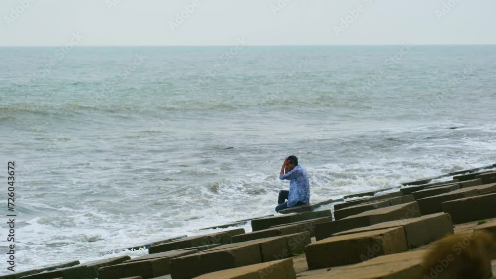 Lonely depressed South Asian Man sitting by the sea in sadness on the slope of the concrete block on the coastline as sea waves hit the coastline. Lonely depressed South Asian Man sitting by the sea in sadness on the slope of the concrete block on the coastline as sea waves hit the coastline.