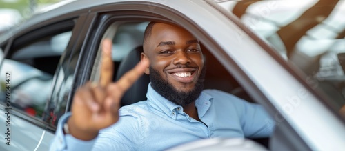 Happy African American male sitting in a new car, showing an okay sign gesture, enjoying his vacation while looking at the camera through an open window in a dealership. photo
