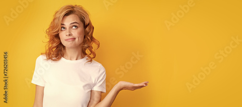 pondering redhead woman presenting product on yellow background with copy space. Beautiful woman isolated face portrait, banner with mock up copy space.
