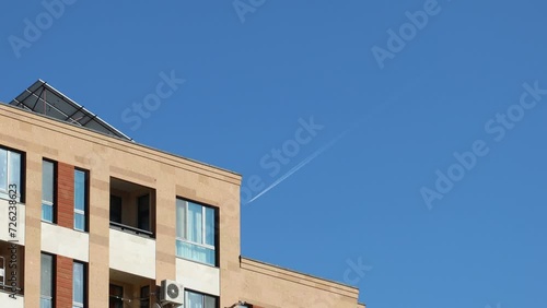 Wallpaper Mural A distant passenger airplane soars high in the clear blue sky, leaving a vapor trail, captured from below, with a building in the foreground Torontodigital.ca