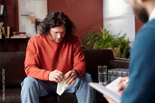 Young man suffering depression holding tissue sitting on couch with head lowered at therapy session