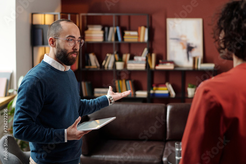 Selective focus medium portrait of Middle Eastern psychologist holding digital tablet welcoming his patient