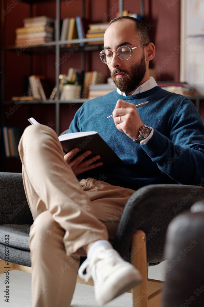Vertical medium long shot of young Middle Eastern psychologist wearing smart casual outfit sitting on chair taking notes in notebook Vertical medium long shot of young Middle Eastern psychologist wearing smart casual outfit sitting on chair taking notes in notebook