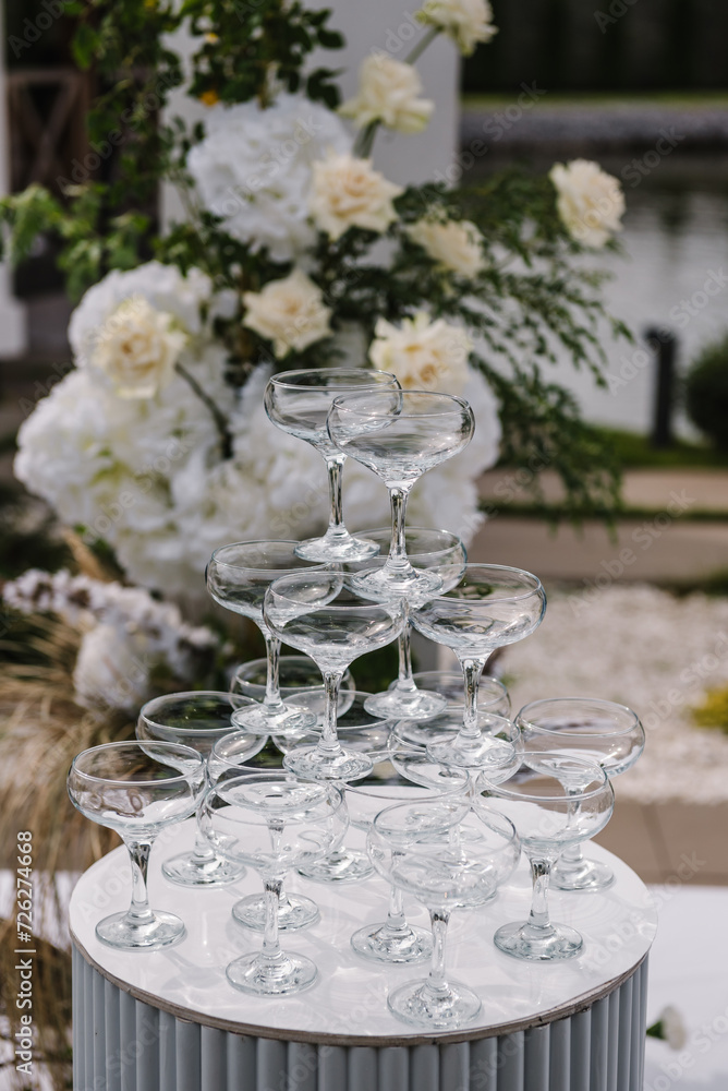 Pyramid of champagne glasses at a party. Wedding decorations in luxury ceremony. Location for celebration on pier. White arch for ceremony is decorated flowers and greenery in backyard with lake view.