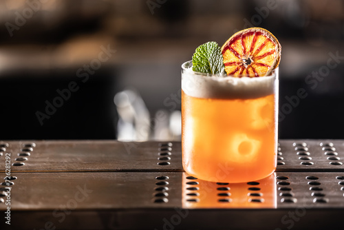 Glass of Jungle Bird cocktail garnished with dried red orange and mint leave on bar counter