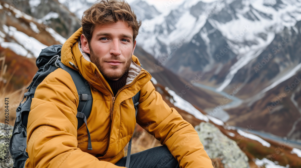 Man wearing vibrant yellow jacket and equipped with a backpack, sits amidst the breathtaking backdrop of snow-capped mountains and winding valleys. Man wearing vibrant yellow jacket and equipped with a backpack, sits amidst the breathtaking backdrop of snow-capped mountains and winding valleys.