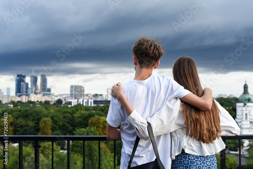 a guy and a girl, hugging each other, stand on the roof of a building, admiring the beautiful view of the city landscape