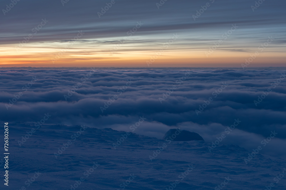 Sunset high in the mountains in winter. White clouds merge with snow to create a soft landscape. A streak of yellow-orange sky can be seen in the distance