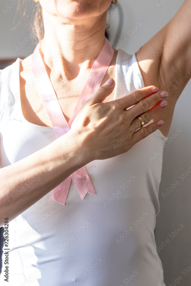 Shot of the woman in the white top, with pink ribbon on her neck as a symbol of breast cancer awareness, performing self examination of the breasts, looking for abnormalities. Concept