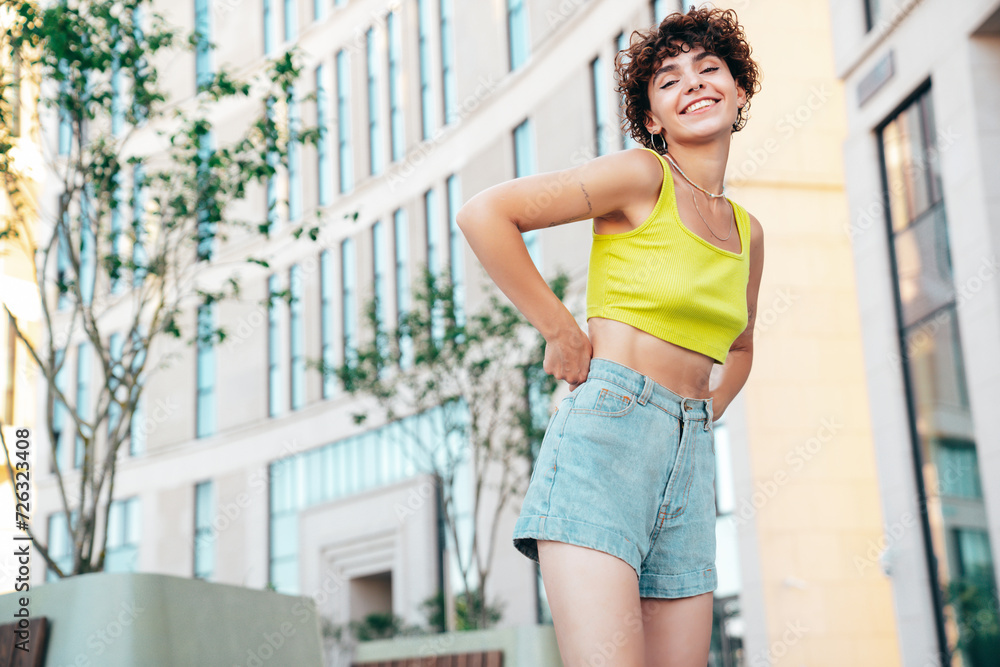Young beautiful smiling hipster woman in trendy summer clothes. Carefree woman with curls hairstyle, posing in the street at sunny day. Positive model outdoors. Cheerful and happy