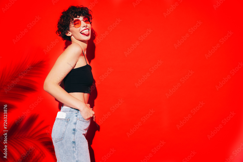 Young beautiful smiling female in trendy summer black tank top and jeans. Carefree woman posing near red wall in studio with curly hairstyle. Positive model having fun. Cheerful and happy. Sunglasses Young beautiful smiling female in trendy summer black tank top and jeans. Carefree woman posing near red wall in studio with curly hairstyle. Positive model having fun. Cheerful and happy. Sunglasses