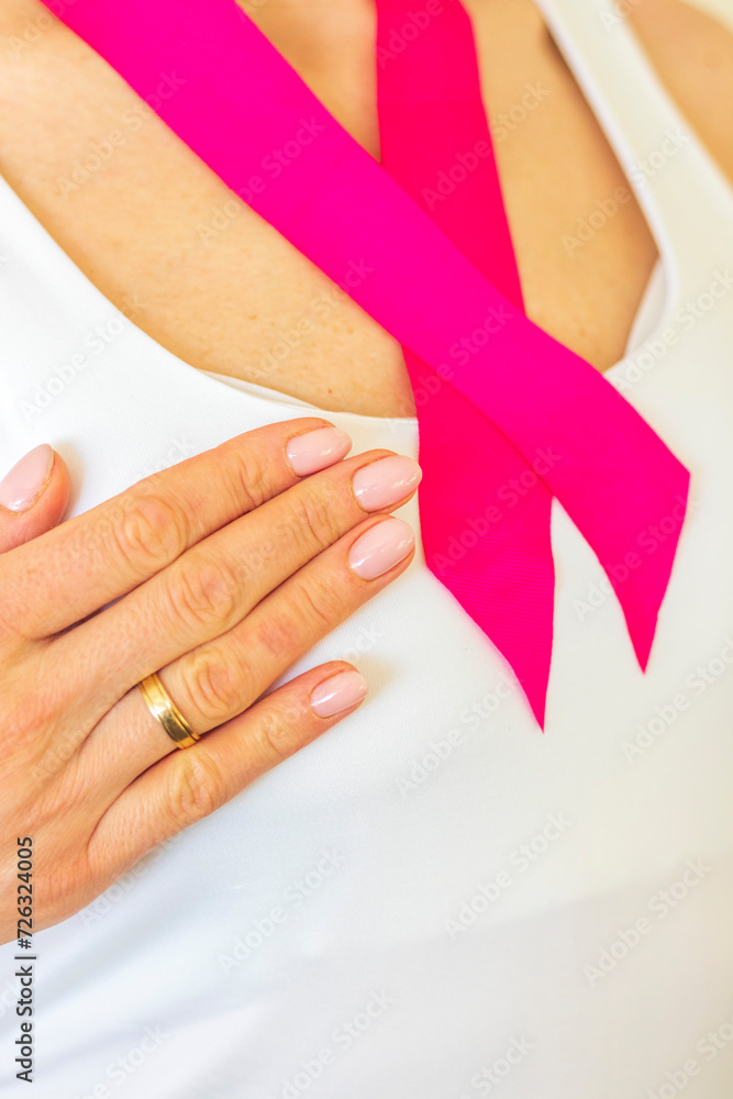 Shot of the woman in the white top against the white wall, with pink ribbon on her neck as a symbol of breast cancer awareness. Concept Shot of the woman in the white top against the white wall, with pink ribbon on her neck as a symbol of breast cancer awareness. Concept