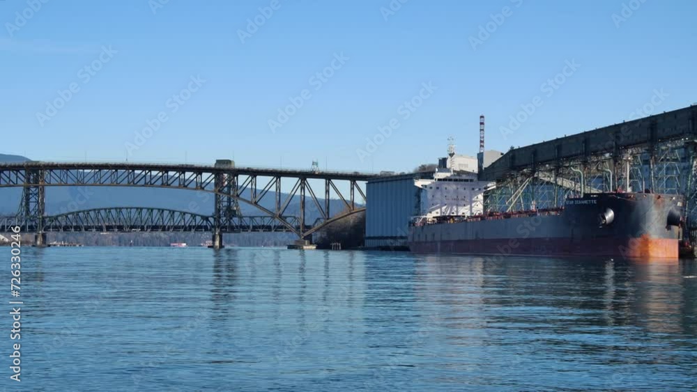 Cargo ship docked at industrial port in Vancouver, sunny day with clear blue sky, bridge in the background, calm waters Cargo ship docked at industrial port in Vancouver, sunny day with clear blue sky, bridge in the background, calm waters