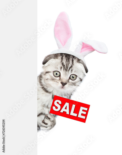 Tabby kitten wearing easter rabbits ears holds sales symbol behind empty white banner. Isolated on white background