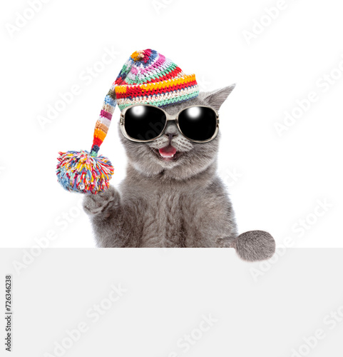 Happy cat wearing sunglasses and knitted warm woolen hat with pompon looks above empty white board. isolated on white background