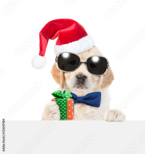 Cute Golden retriever puppy wearing sunglasses, red santa hat and tie bow looks above  empty white banner and holds gift box. isolated on white background