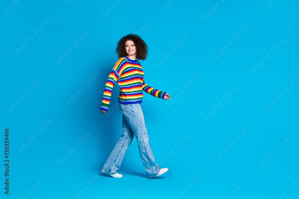 Full size photo of pretty young girl walk turn around look empty space wear trendy rainbow print outfit isolated on blue color background Full size photo of pretty young girl walk turn around look empty space wear trendy rainbow print outfit isolated on blue color background