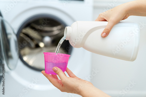 Person adding liquid laundry detergent to the washer, close up. Female hand holding laundry detergent in front of open washing machine