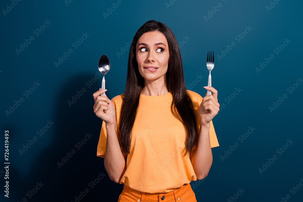 Portrait of biting lips woman wear holding metal spoon and fork looking empty space choose dish isolated on dark blue color background Portrait of biting lips woman wear holding metal spoon and fork looking empty space choose dish isolated on dark blue color background
