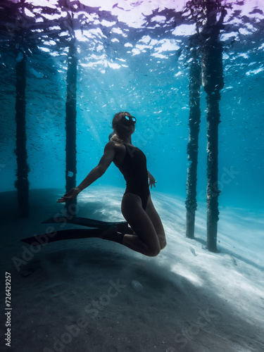 Freediver woman posing underwater under the pier in ocean. Female swims with fins under the pier