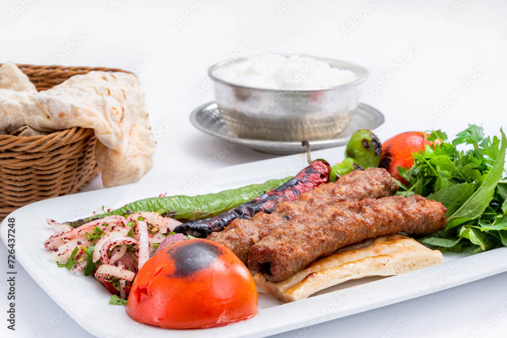 Top view of traditional grilled Adana kebab with cooked spicy shish kebab, pepper, tomato, and fresh greenery on a white plate with a metal pot of foamy ayran and lavash isolated on white background. Top view of traditional grilled Adana kebab with cooked spicy shish kebab, pepper, tomato, and fresh greenery on a white plate with a metal pot of foamy ayran and lavash isolated on white background.