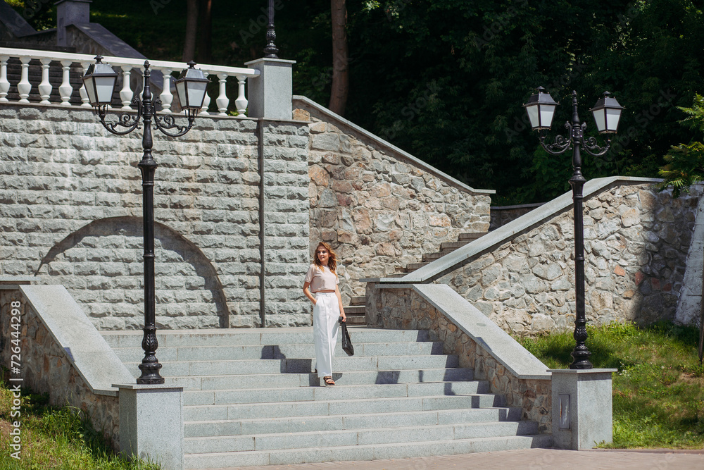 A stylish girl with fashionable makeup and hairstyle, in white trousers and a beige top and a handbag in her hands, stands against the backdrop of a large stone staircase in the park.