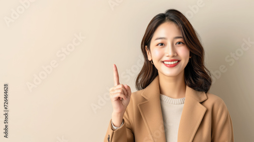 Portrait of a Young Korean woman in a beige coat gesture with the index finger pointing upwards isolated with copy space. photo