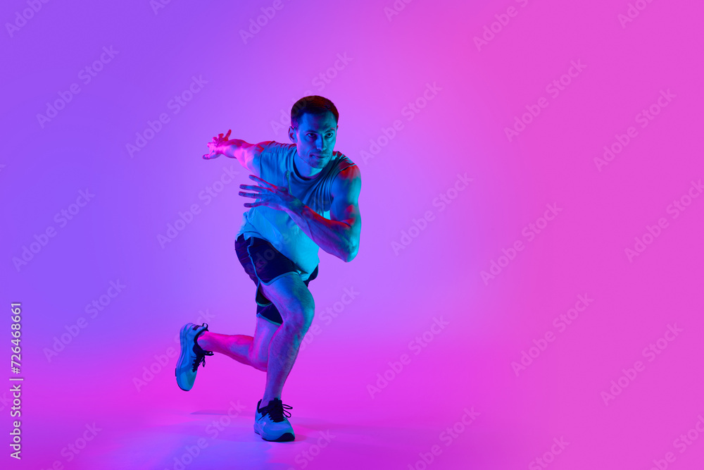 Warming-up exercises. Sportive young man in sportswear training, doing exercises against gradient pink background in neon light. Concept of active and healthy lifestyle, sport, fitness, endurance