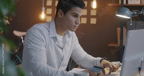 Slow motion of Arab man using credit card and computer celebrating business achievement in dark office in evening. Banking and finance concept. photo