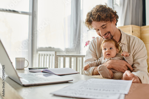 happy father holding infant son while working from home near papers and gadgets, work-life balance photo