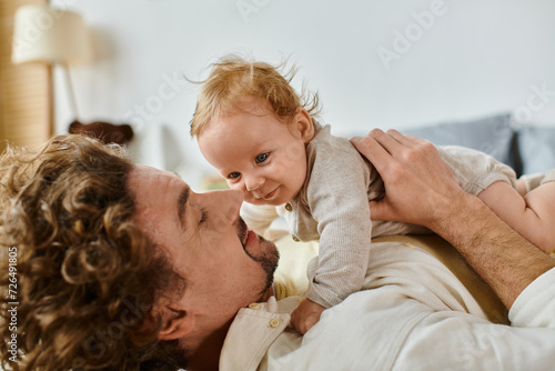 happy man with curly hair and beard embracing with his infant son in bedroom, love and care