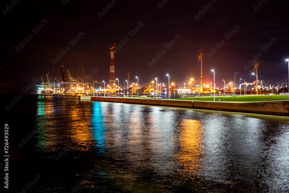 An engaging night scene unfolds at the Port of Antwerp, where the artificial lights create a vibrant tapestry against the dark sky. The reflection of the lights on the water adds a layer of depth and