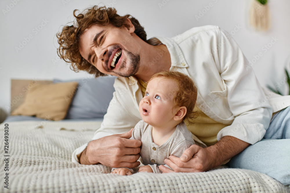 happy man with curly hair laughing while embracing his baby boy and lying together on bed happy man with curly hair laughing while embracing his baby boy and lying together on bed