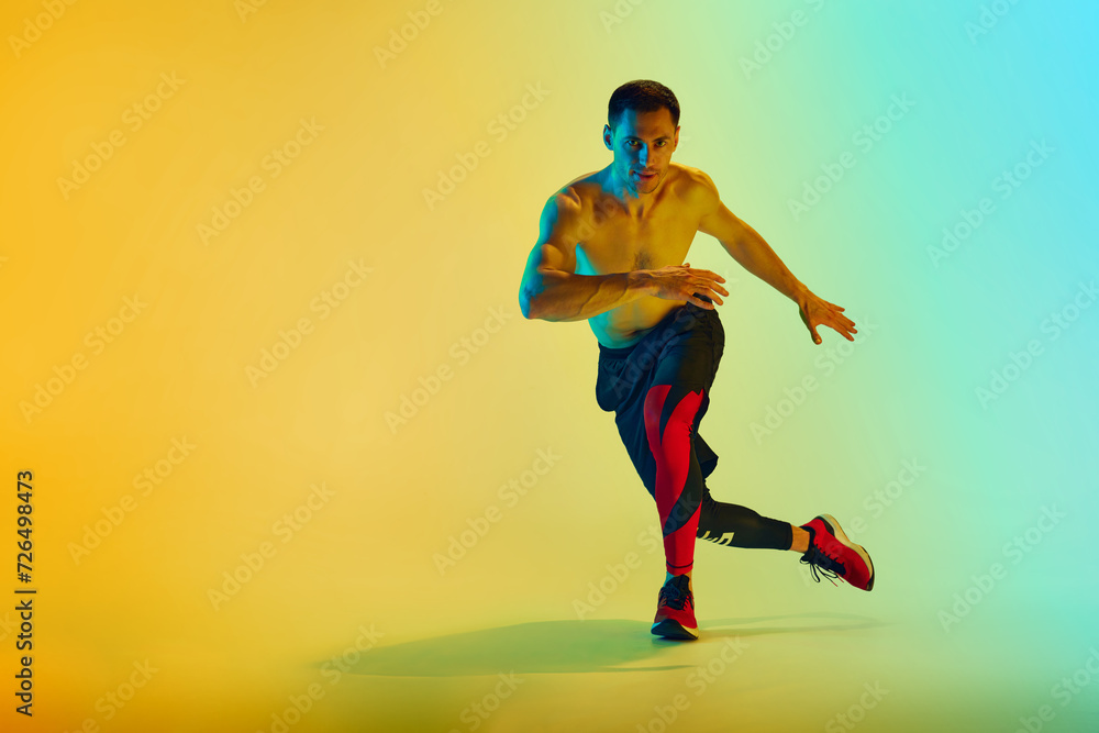 Fototapeta premium Shirtless male athlete in mid-action with focused expression training shirtless against gradient blue yellow background in neon. Concept of active and healthy lifestyle, sport, fitness, endurance
